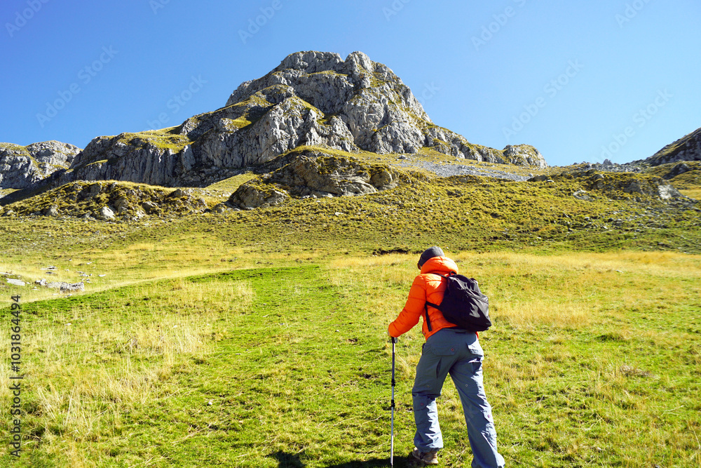 A tourist begins his ascent to the top: a man with a backpack walks along the mountainside using hiking poles. Active recreation in Montenegro in autumn: trekking in the mountains near Kapetanovo Lake A tourist begins his ascent to the top: a man with a backpack walks along the mountainside using hiking poles. Active recreation in Montenegro in autumn: trekking in the mountains near Kapetanovo Lake