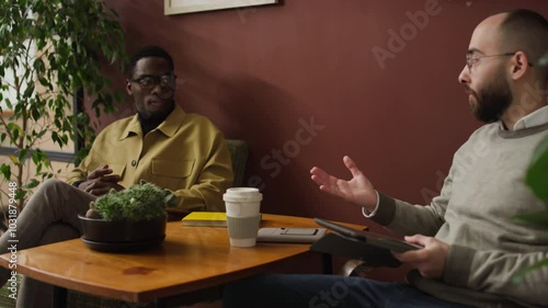 Two diverse young male colleagues discussing business strategy while relaxing in armchairs near coffee table during workday photo