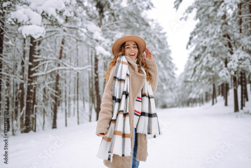 Happy woman among snowy trees in winter forest, enjoying the first snow in hat and coat. Boho style. Active lifestyle and tourism.