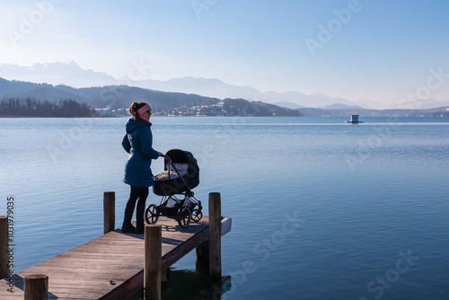 Woman with baby stroller standing on pier at idyllic Lake Wörth in Pörtschach, Carinthia, Austria. Scenic landscape surrounded by Karawanks, Austrian Alps. Fresh and clean air. Calm sunny winter day