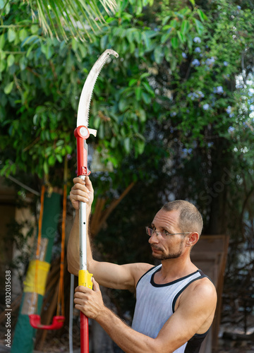  Man outdoors holding a long pruning saw. He is wearing glasses and a t-shirt, focusing on adjusting the pole saw. The background includes lush greenery, garden. The garden work, tree maintenance