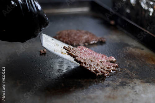 Hand using a spatula to flip cooking smash burgers on a grill in a restaurant kitchen