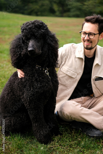 Young man on the park lawn playing with a royal black poodle