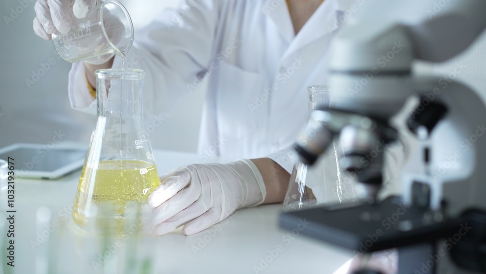 A scientist, wearing a lab coat and white protective gloves, is pouring a yellow oily liquid from one beaker to another near microscope in laboratory, close up. Medicine and science concept A scientist, wearing a lab coat and white protective gloves, is pouring a yellow oily liquid from one beaker to another near microscope in laboratory, close up. Medicine and science concept