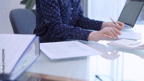 A female accountant with blue dotted blousy is using a laptop computer and calculator to calculate taxes at a glass desk in the office. Right side view photo