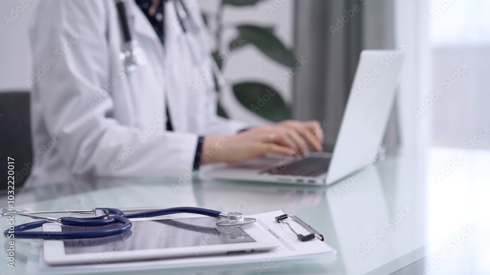 Tablet computer and blue stethoscope are on the glass table while doctor woman is using laptop computer on the glass table. Medicine concept Tablet computer and blue stethoscope are on the glass table while doctor woman is using laptop computer on the glass table. Medicine concept