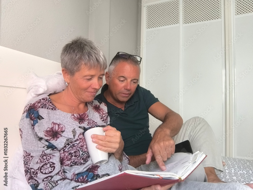 Young couple in bed interested in reading a book.Boy and girl reading a book with passion.Caucasian couple in bed relaxed with a book and talking.she with cup in hand looks at the book he points out. Young couple in bed interested in reading a book.Boy and girl reading a book with passion.Caucasian couple in bed relaxed with a book and talking.she with cup in hand looks at the book he points out.