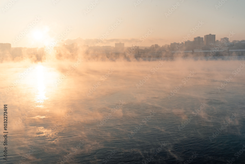 Cold fog over the river at the sunset with city on the background.