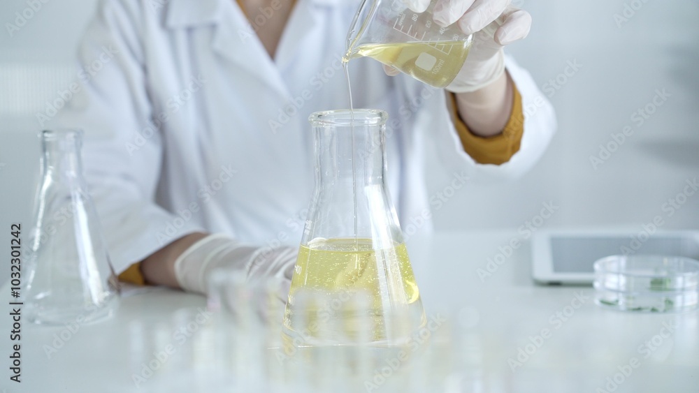 A scientist, wearing a lab coat and white protective gloves, is pouring a yellow oily liquid from one beaker to another in laboratory, close up. Medicine and science concept A scientist, wearing a lab coat and white protective gloves, is pouring a yellow oily liquid from one beaker to another in laboratory, close up. Medicine and science concept