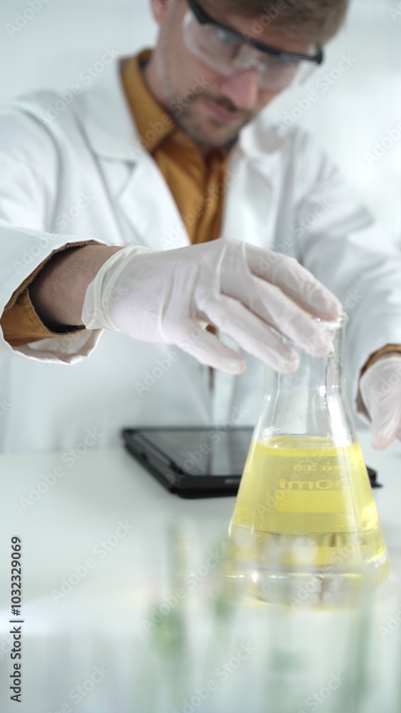 Man scientist wearing white protective gloves, and glasses is holding a yellow chemical solution inside an Erlenmeyer flask in a laboratory, vertical portrait Man scientist wearing white protective gloves, and glasses is holding a yellow chemical solution inside an Erlenmeyer flask in a laboratory, vertical portrait