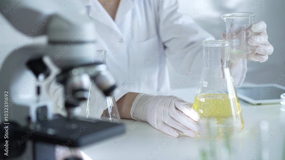 Fototapeta premium A scientist, wearing a lab coat and white protective gloves, is pouring a yellow oily liquid from one beaker to another near microscope in laboratory, close up. Medicine and science concept