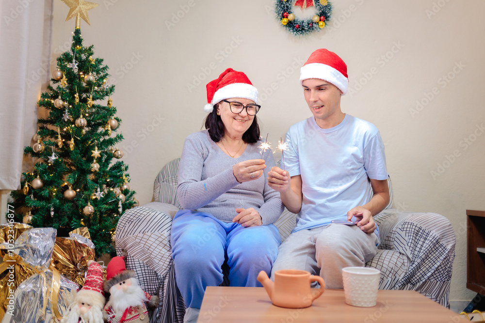 A Brazilian mother and adult son celebrating Christmas at home. They are wearing Santa hats and are happy as they hold lit sparkler candles to celebrate in a festively decorated room A Brazilian mother and adult son celebrating Christmas at home. They are wearing Santa hats and are happy as they hold lit sparkler candles to celebrate in a festively decorated room