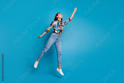 Full size photo of gorgeous girl dressed print blouse jeans pants flying catch object look empty space isolated on blue color background