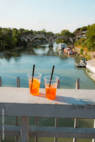 Wallpaper Mural Two Aperol spritz drinks on a riverside ledge with a scenic view of a historic stone bridge in Rome. The refreshing cocktails are set against a picturesque backdrop of the Tiber River and greenery. Torontodigital.ca