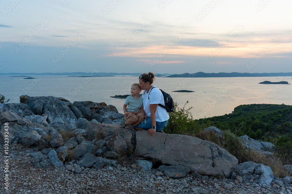 Mother and young son enjoying sunset on rocky hilltop with scenic sea and island view. Concept of family bonding, outdoor adventure, and nature appreciation. Mother and young son enjoying sunset on rocky hilltop with scenic sea and island view. Concept of family bonding, outdoor adventure, and nature appreciation.
