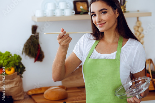 An attractive young dark-haired woman tasting ready hot meal with a wooden spoon while standing and smiling near the kitchen stove. Cooking and householding concepts