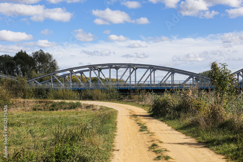 Metal bridge background. Road bridge construction. Empty asphalt road. Blue sky weather long bridge over Narew river. Tykocin village in Poland. Steel structure landscape. Wild nature along the river.