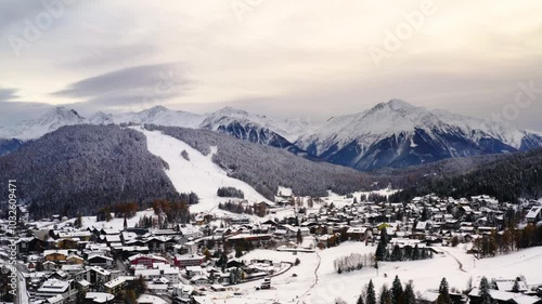 Ski mountain covered with white snow in the alps next to alpine village filled by ski chalet shot from arial drone