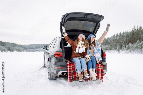 Happy friends in winter forest in the car. Two happy girls sit in the trunk of a car talking and laughing.