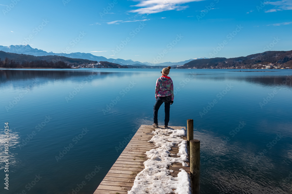 Tourist woman standing on pier with view of idyllic Lake Wörth in Pörtschach, Carinthia, Austria. Scenic landscape surrounded by Karawanks, Austrian Alps. Fresh and clean air. Calm sunny winter day Tourist woman standing on pier with view of idyllic Lake Wörth in Pörtschach, Carinthia, Austria. Scenic landscape surrounded by Karawanks, Austrian Alps. Fresh and clean air. Calm sunny winter day
