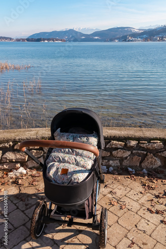 Baby stroller standing at lakeshore of idyllic Lake Wörth in Pörtschach, Carinthia, Austria. Scenic landscape surrounded by Karawanks, Austrian Alps. Happy family walk on relaxed sunny winter day photo