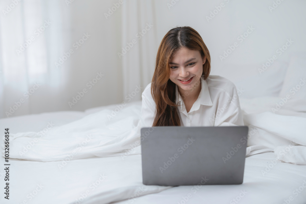 Young woman is using a laptop computer while lying on her bed, working remotely from home Young woman is using a laptop computer while lying on her bed, working remotely from home