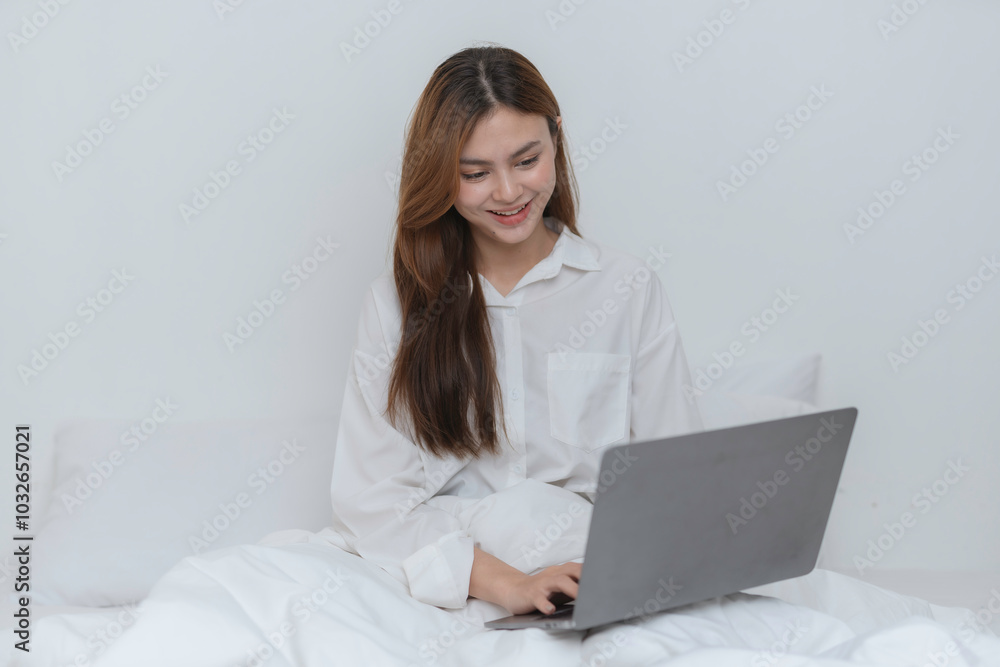 Smiling young woman with long brown hair wearing a white shirt working on a laptop while sitting in bed Smiling young woman with long brown hair wearing a white shirt working on a laptop while sitting in bed