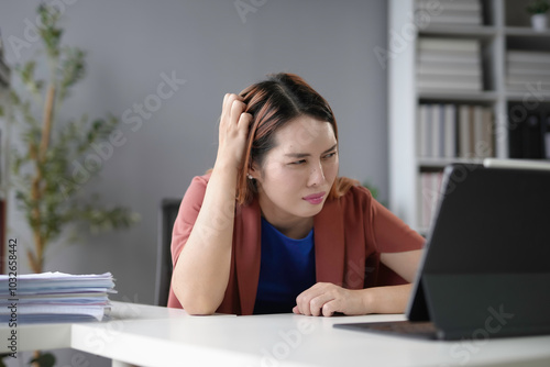 Young asian businesswoman is scratching her head with a confused expression while looking at a tablet in her office