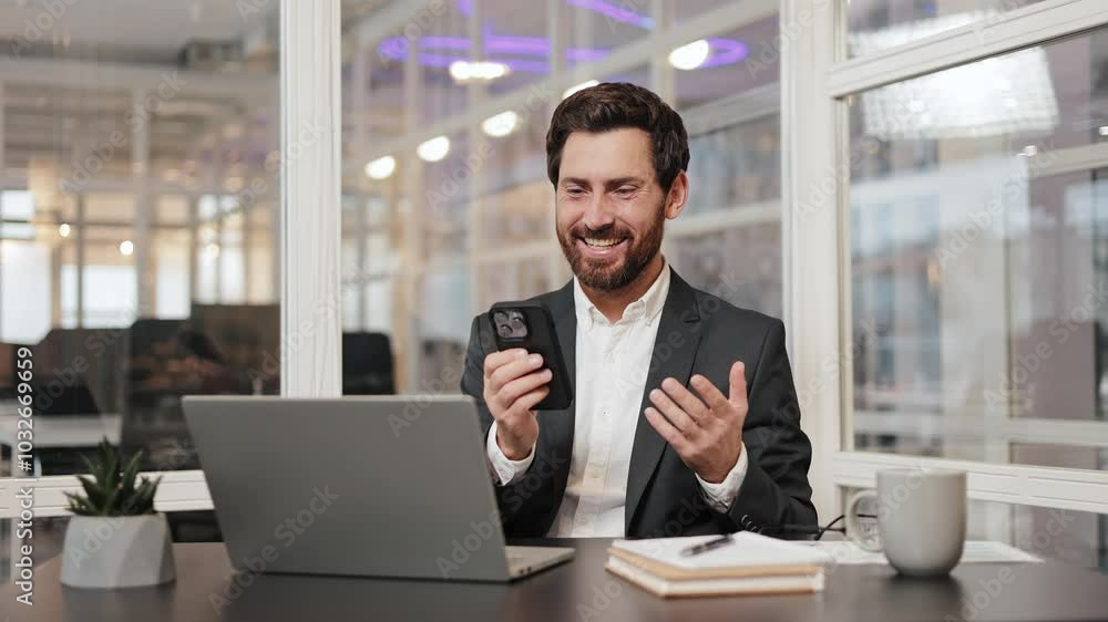 A happy businessman in a suit is sitting at his desk in the office. He looks at his phone and smiles. He is successful. He is glad to receive good news. A happy businessman in a suit is sitting at his desk in the office. He looks at his phone and smiles. He is successful. He is glad to receive good news.