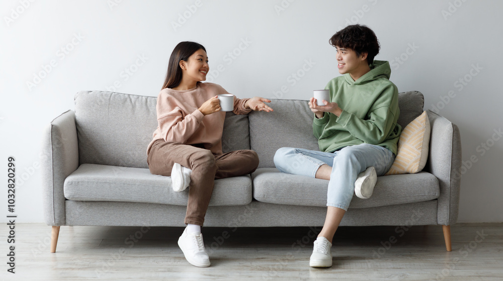 Young Asian man and woman drinking coffee and talking while sitting on couch at home. Loving millennial couple enjoying coffee while having conversation, spending weekend together Young Asian man and woman drinking coffee and talking while sitting on couch at home. Loving millennial couple enjoying coffee while having conversation, spending weekend together