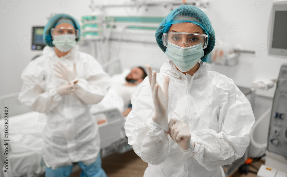 Two healthcare workers in full protective gear, one adjusting gloves, stand in hospital room with a male patient in the background, emphasizing readiness, safety, and professionalism in patient care Two healthcare workers in full protective gear, one adjusting gloves, stand in hospital room with a male patient in the background, emphasizing readiness, safety, and professionalism in patient care