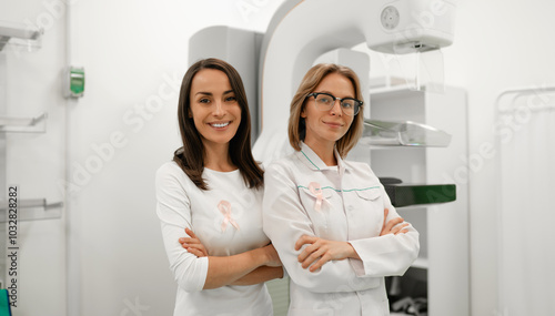 Two women, doctor and patient, smile confidently in front of a mammography machine. Both wear pink breast cancer awareness ribbons, promoting early detection and women's health in a medical facility