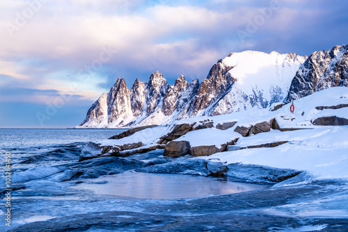 Tungeneset or Devils Jaw mountain on Senja island, Norway in winter. Winter landscape with snowy mountain peaks, fjord waters and ice coast in northern Norway. Scenic view with dramatic sky