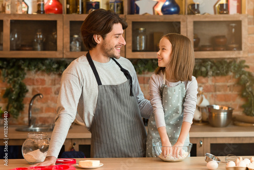 Happy young father and little daughter baking pie in kitchen together, kneading dough, cooking at home, enjoying homemade pastry, wearing aprons and smiling to each other, copy space