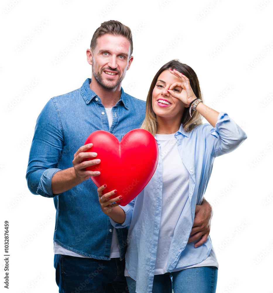 Young couple in love holding red heart over isolated background with happy face smiling doing ok sign with hand on eye looking through fingers Young couple in love holding red heart over isolated background with happy face smiling doing ok sign with hand on eye looking through fingers