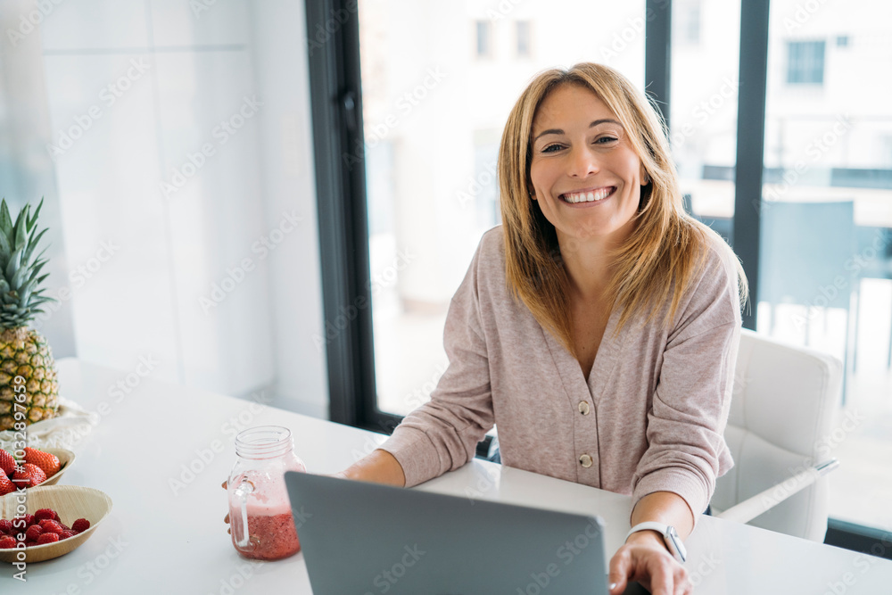 Smiling Woman Holding Smoothie by Laptop in Bright Kitchen