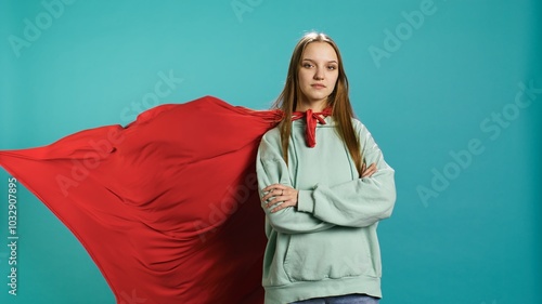 Happy young girl portraying superhero flying with cape fluttering in wind, isolated over studio background. Portrait of upbeat heroic woman posing as hero in costume, camera B