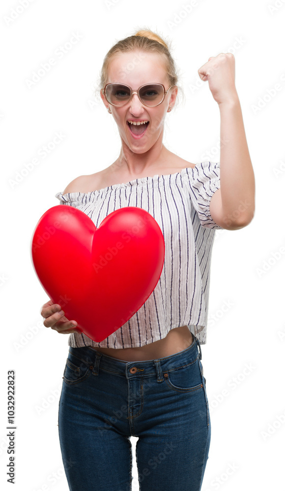 Blonde teenager woman holding red heart annoyed and frustrated shouting with anger, crazy and yelling with raised hand, anger concept Blonde teenager woman holding red heart annoyed and frustrated shouting with anger, crazy and yelling with raised hand, anger concept