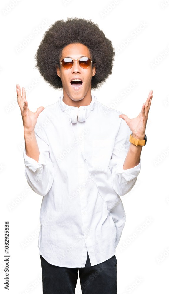 Young african american man with afro hair wearing sunglasses and headphones crazy and mad shouting and yelling with aggressive expression and arms raised. Frustration concept. Young african american man with afro hair wearing sunglasses and headphones crazy and mad shouting and yelling with aggressive expression and arms raised. Frustration concept.