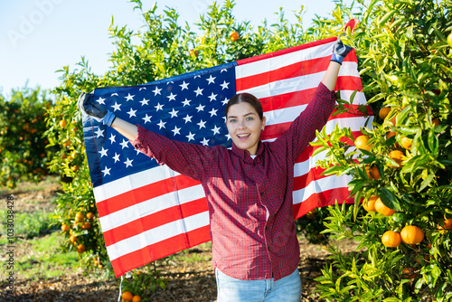 Portrait of a young confident farmer woman in a fruit nursery, holding the flag of USA in her hands and demonstrating it photo