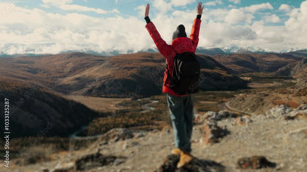 A female tourist in an orange jacket with a backpack on her shoulders goes to the top of the mountain and rejoices at her achievement by raising his hands up. Travel and tourism concept. A female tourist in an orange jacket with a backpack on her shoulders goes to the top of the mountain and rejoices at her achievement by raising his hands up. Travel and tourism concept.