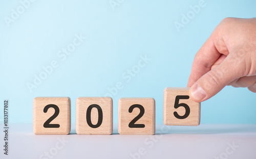 A hand adjusts wooden blocks with numbers to form the year 2025 against a light blue background, symbolizing new beginnings and future goals