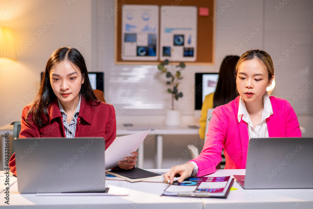 Two women are sitting at a desk with laptops and papers