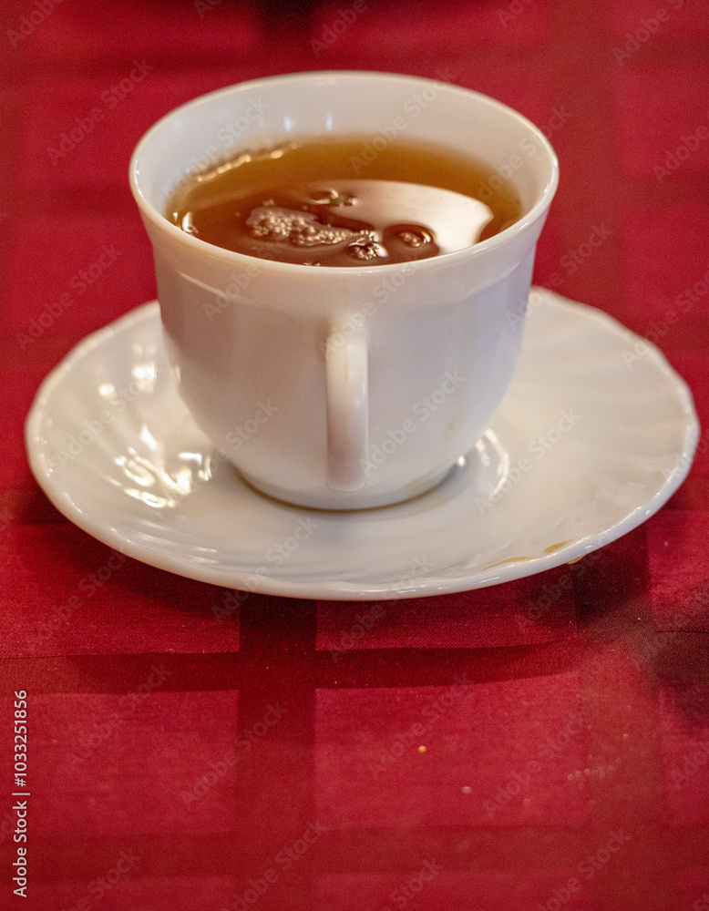 A white cup of tea sits on a white saucer on a red tablecloth A white cup of tea sits on a white saucer on a red tablecloth