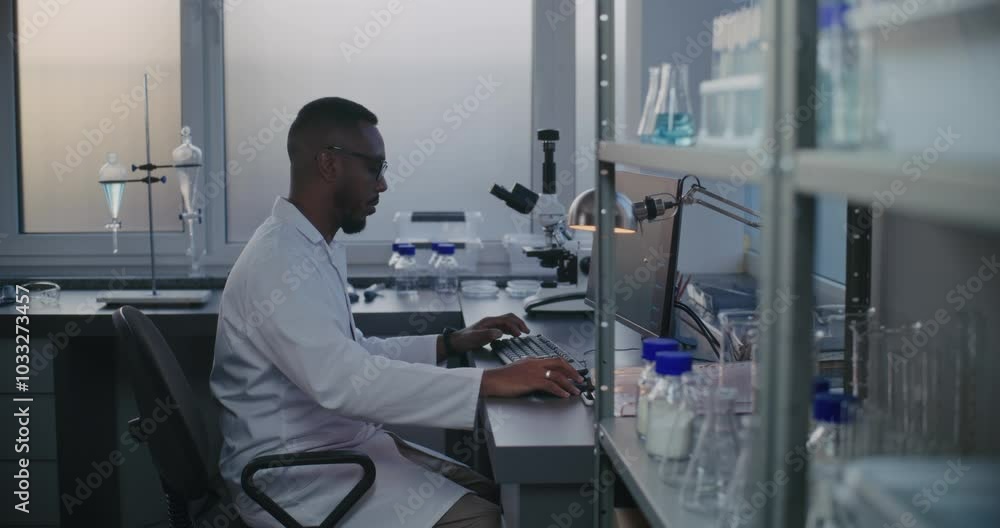 Medical science lab with African American professional biotechnology scientist working on personal computer, typing on keyboard. Advanced equipment in chemical laboratory: PC, microscope, glassware. Medical science lab with African American professional biotechnology scientist working on personal computer, typing on keyboard. Advanced equipment in chemical laboratory: PC, microscope, glassware.
