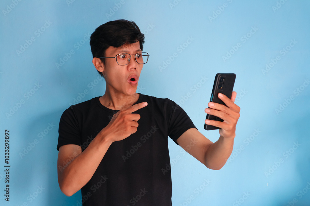 Asian young man looking at his mobile phone with amazed expression Asian young man looking at his mobile phone with amazed expression