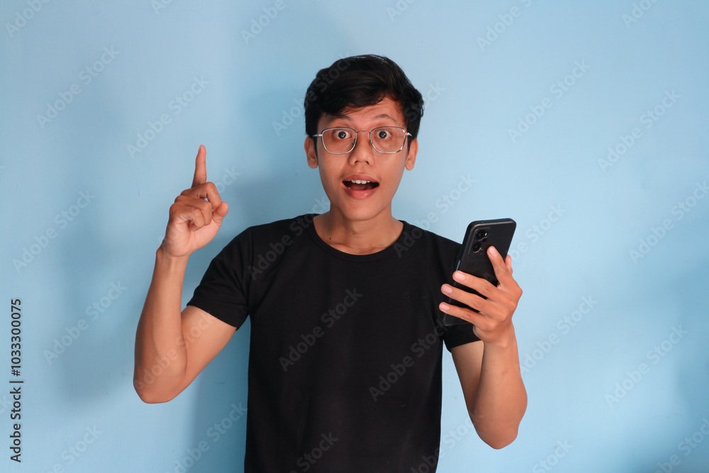 Young Asian man holding a cell phone while pointing upwards with a happy expression Young Asian man holding a cell phone while pointing upwards with a happy expression