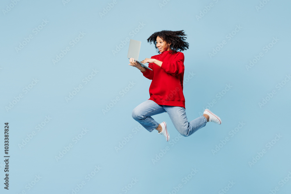 Full body little kid teen IT girl of African American ethnicity wear red sweatshirt jump high hold use work on laptop pc computer isolated on pastel plain blue background. Childhood lifestyle concept. Full body little kid teen IT girl of African American ethnicity wear red sweatshirt jump high hold use work on laptop pc computer isolated on pastel plain blue background. Childhood lifestyle concept.