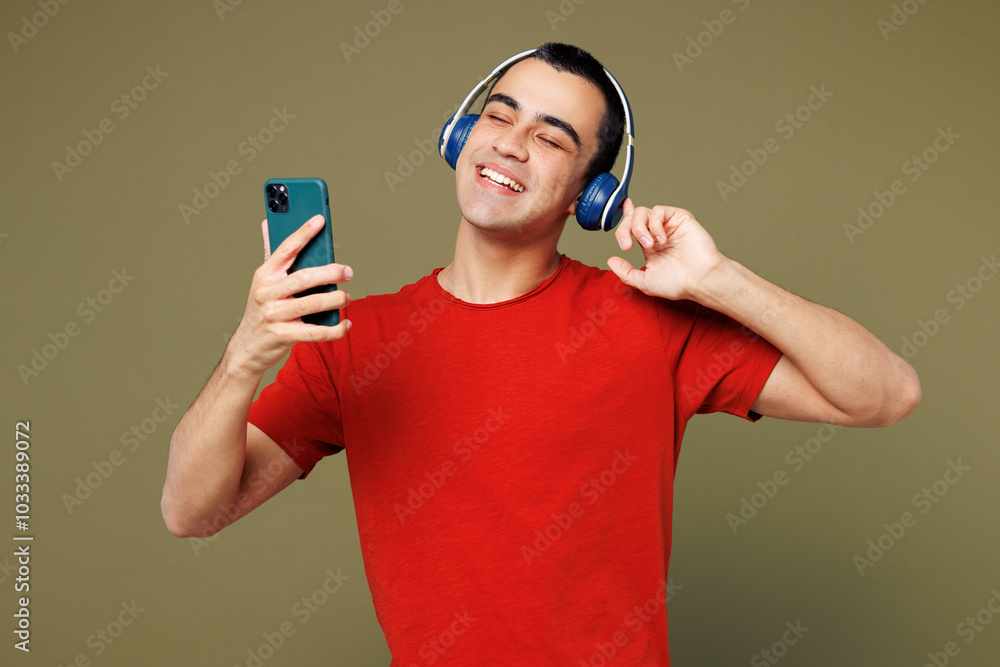 Young middle eastern man he wear red t-shirt casual clothes listen to music in headphones use hold mobile cell phone enjoy isolated on plain pastel green background studio portrait. Lifestyle concept. Young middle eastern man he wear red t-shirt casual clothes listen to music in headphones use hold mobile cell phone enjoy isolated on plain pastel green background studio portrait. Lifestyle concept.