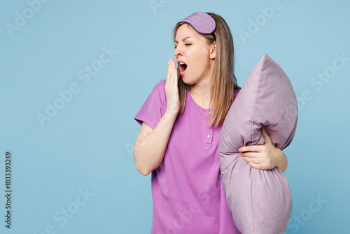 Calm sad young woman wear purple pyjamas jam sleep eye mask rest relax at home yaw cover mouth with hand hold pillow isolated on plain pastel light blue cyan background. Good mood night nap concept.